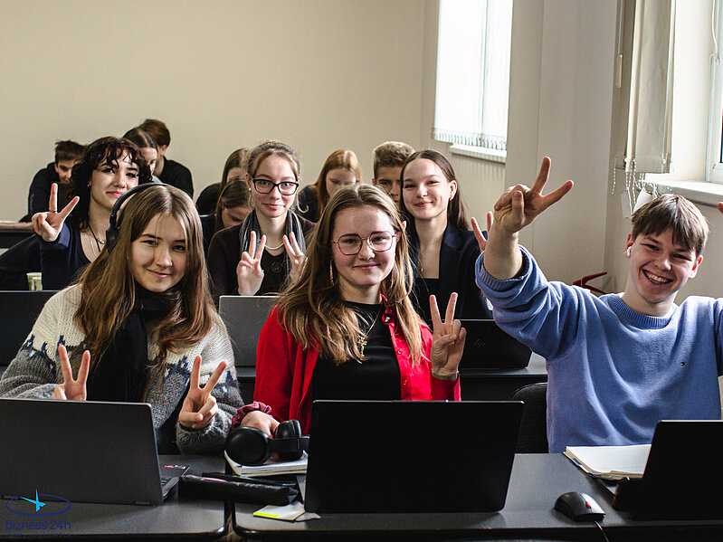 Konkursa dalībnieki pozē fotogrāfijai lekciju telpā, smaida un rāda  žestus ar rokām. Fonā redzami vēl citi dalībnieki un klēpjdatori uz soliem.