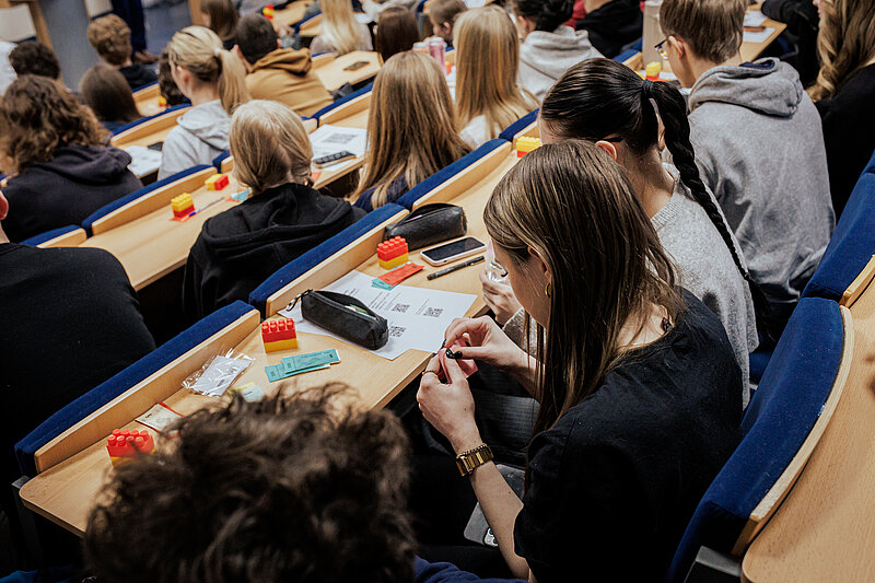 Skats no augšas uz studentiem, kas sēž lekciju zālē un pilda konkursa uzdevumu. Redzami vairāki klēpjdatori un papīri uz soliem, studenti ir iegrimuši darbā. Viņiem rokās ir Lego klucīši.