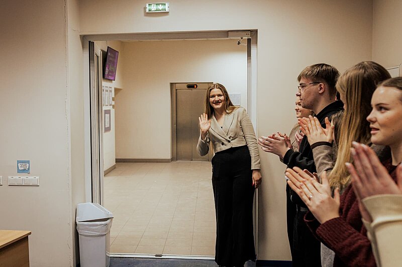 Konkursa dalībnieki aplaudē. Durvju ailē stāv studente un māj sveicienu.