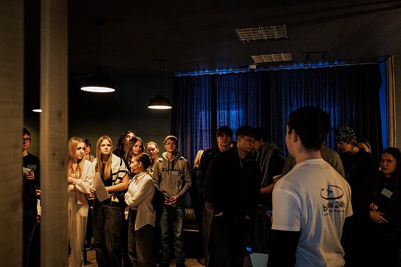 Studentu grupa pulcējas tumšā telpā ar zilganu apgaismojumu fonā. Dalībnieki sarunājas mazākās grupās.