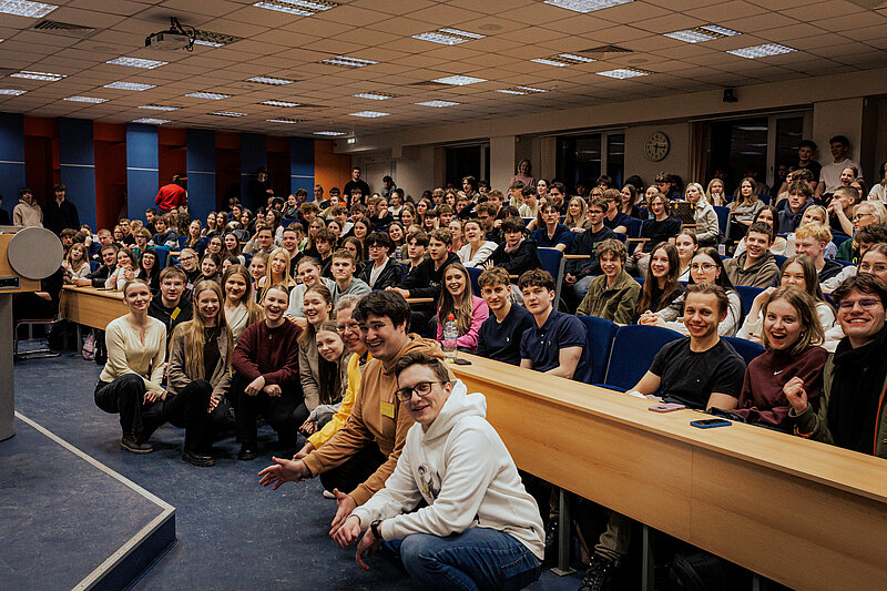 Liela auditorija ar daudziem konkursa dalībniekiem, kas sēž lekciju zālē un skatās uz priekšu. Zāle ir  pilna, redzami daudzi jauni cilvēki,  studenti un skolēni.
